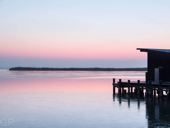 Holzsteg führt ins ruhige Gewässer bei Sonnenuntergang mit weichem rosa Himmel, der sich im Wasser spiegelt.